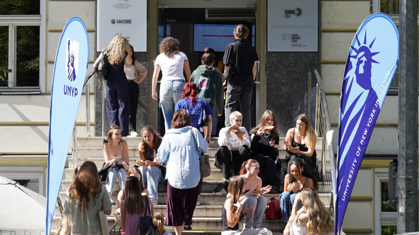 Students sitting and walking on the front steps of the University of New York in Prague building, with UNYP-branded banners on both sides of the entrance.